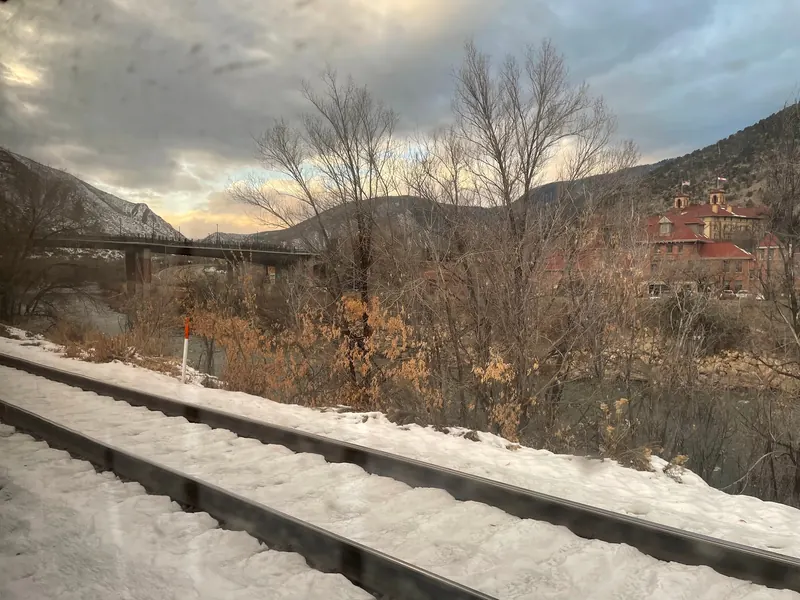 Snowy railway foreground with a river bridge and Glenwood Hot Springs building beyond; not a recommended walking route or river swimming access