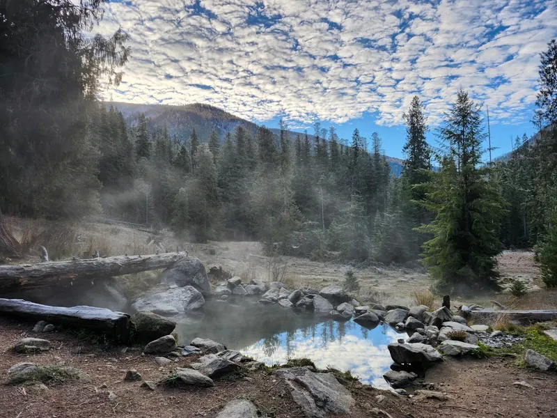 Rock-bordered pool, drifting mist and evergreen forest in the Jerry Johnson Hot Springs collection; historical view, not a current temperature or water-quality report