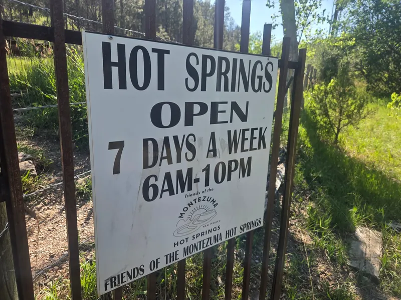 Historical Friends of Montezuma Hot Springs opening-hours sign beside a wooden fence