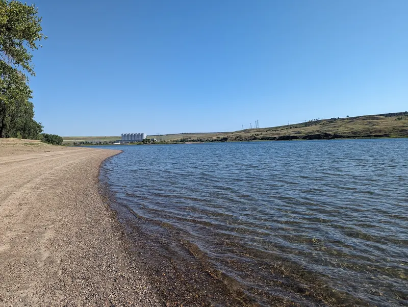 Empty sandy shoreline with blue water and a distant dam structure in the Oahe Downstream collection, not a verified swim entrance
