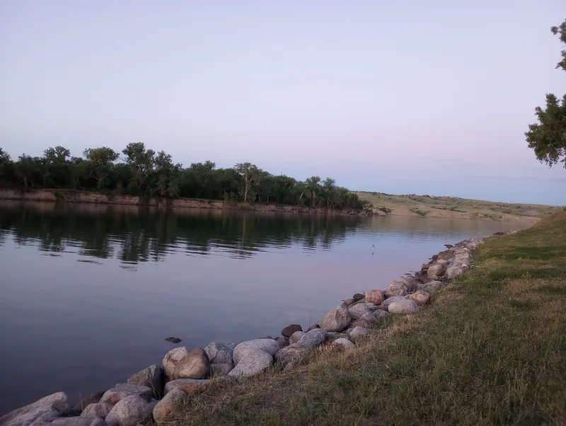 Rock-lined grassy shoreline beside reflective water and distant trees at dusk in the Oahe Downstream collection