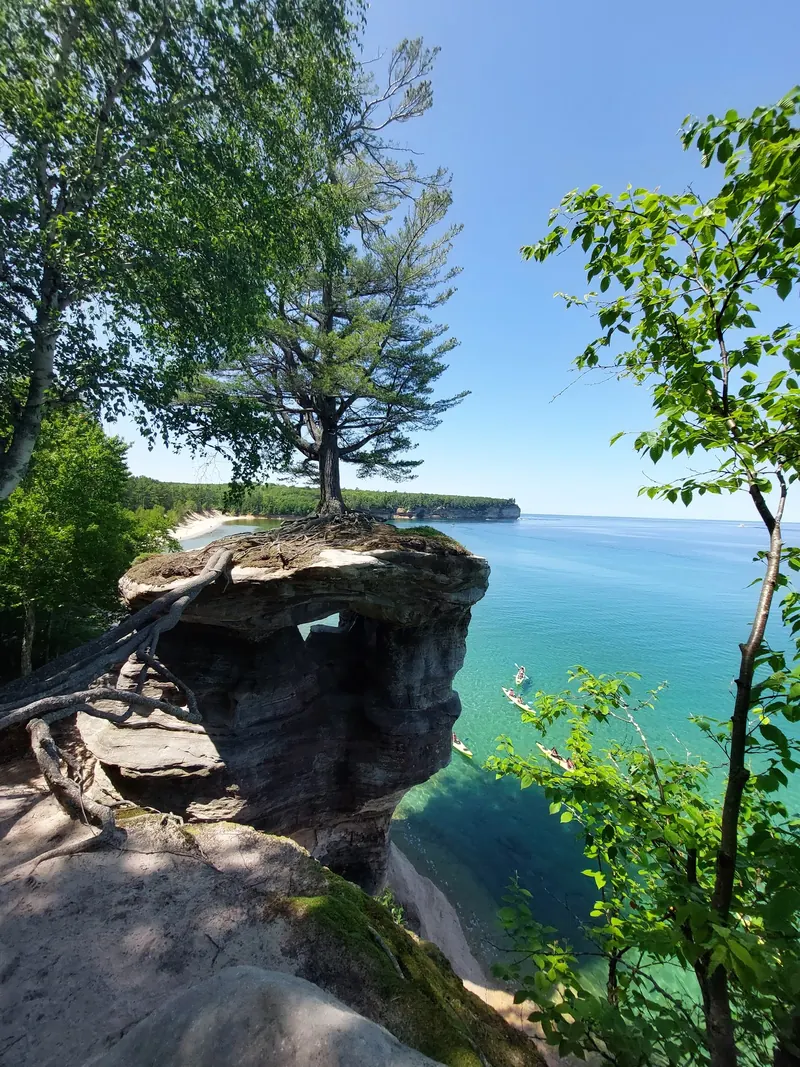 Exposed tree roots above a lakeside rock arch with distant kayakers in the Pictured Rocks collection