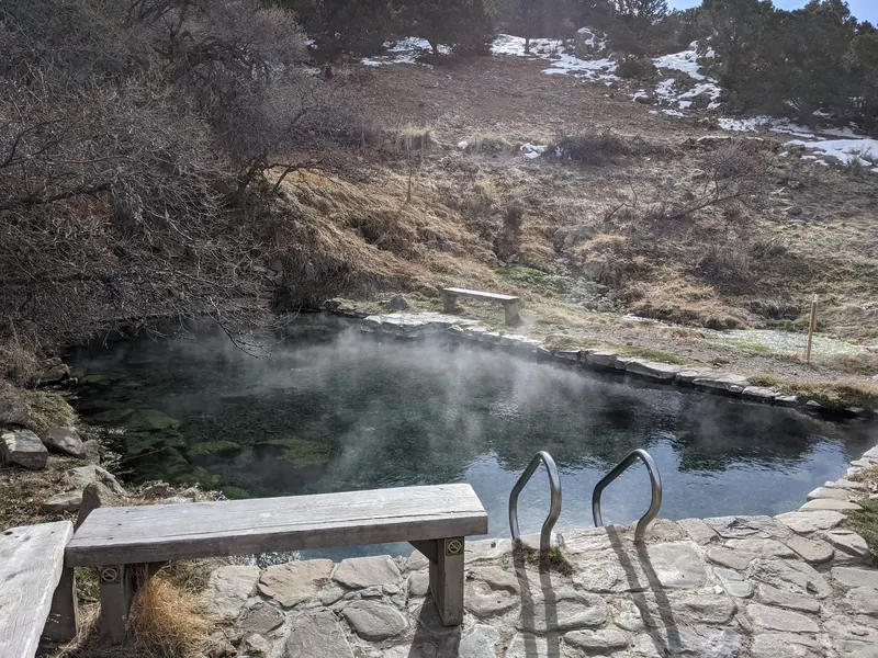 Empty steaming rock-lined pool with handrails and a bench at Valley View Hot Springs