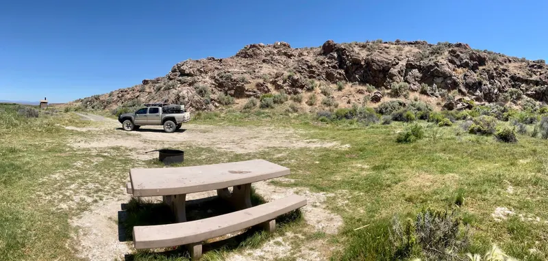 Picnic table beside a rocky bluff with a parked pickup and distant toilet structure in the Willow Creek Hot Springs collection