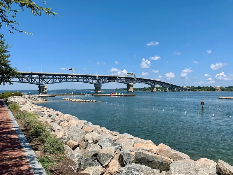 Rock-lined Yorktown waterfront beside a brick walkway, with a buoy line, docks and bridge; historical shoreline context, not current swimming clearance